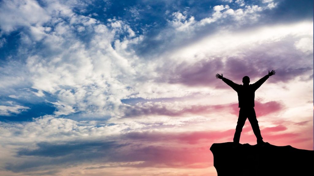 Celebrating freedom; person on cliff throwing up hands in joy at sunrise.